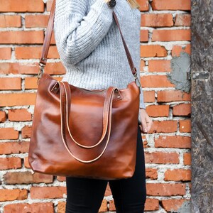 May include: A large, cognac-brown leather tote bag featuring two sturdy top handles and a detachable, adjustable shoulder strap with antique brass-toned metal hardware. The bag has a zippered top closure and a smooth, polished finish.