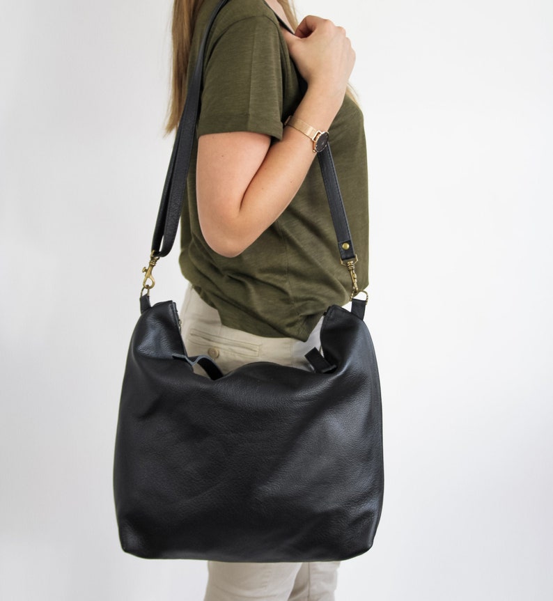 May include: A black leather hobo bag with a soft, slouchy silhouette and a wide shoulder strap. The bag features antique brass-toned metal hardware, including swivel clips that attach the strap to the sides of the bag.