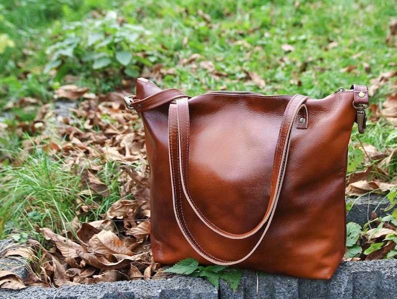 May include: A medium-sized brown leather tote bag with a smooth, polished finish. The bag features two long, thin shoulder straps, a top zipper closure, and metallic hardware attachments on the sides for an adjustable strap.