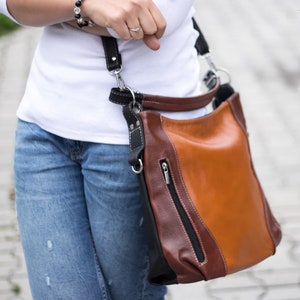 Puede incluir: Un bolso de hombro de cuero marr&oacute;n y beige con una correa negra. El bolso tiene un bolsillo con cremallera en la parte delantera.