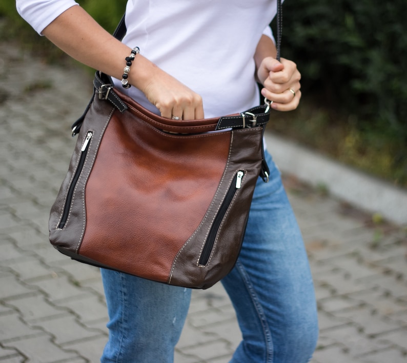Pode incluir: Uma bolsa de ombro de couro marrom com detalhes pretos e z&iacute;peres. A bolsa tem uma al&ccedil;a longa e &eacute; usada no ombro.