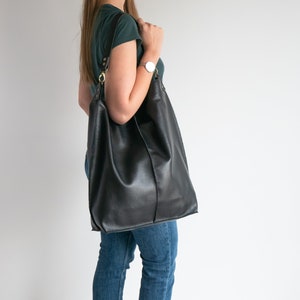 K&ouml;nnte beinhalten: Eine schwarze Ledertasche mit goldener Schnalle und Trageriemen. Die Tasche wird von einer Person getragen, die ein gr&uuml;nes Shirt und eine blaue Jeans tr&auml;gt.