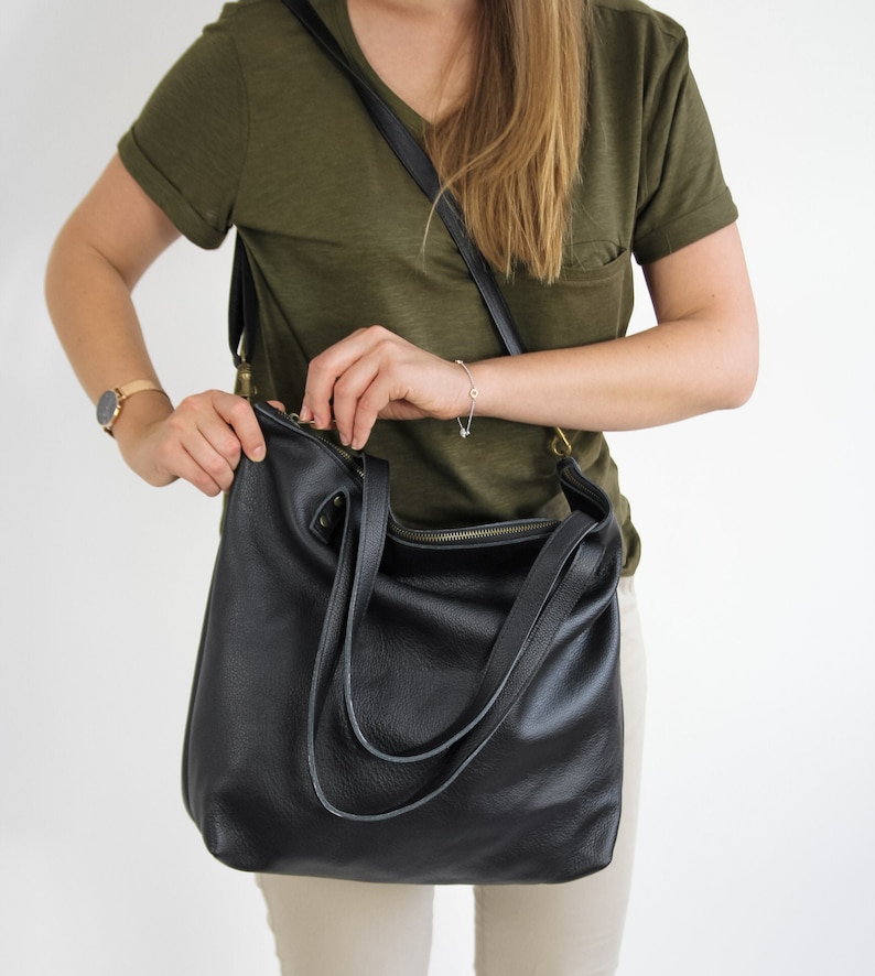 May include: A black leather hobo bag featuring a top zipper closure, two shoulder straps, and a detachable crossbody strap with brass-toned hardware. The bag has a pebbled texture and a slouchy, unstructured shape with visible stitching details.