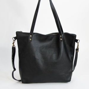 May include: A black leather tote bag featuring two long shoulder straps attached with brass-colored rivets, a top zipper closure, and a detachable shoulder strap connected to the sides with brass-colored metal swivel clasps.