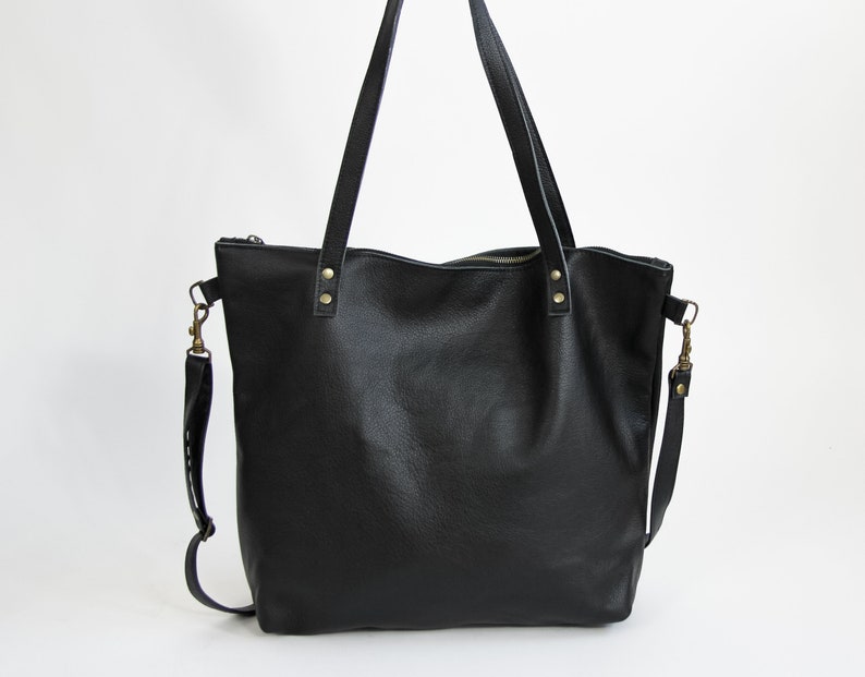 May include: A black leather tote bag featuring two long shoulder straps attached with brass-colored rivets, a top zipper closure, and a detachable shoulder strap connected to the sides with brass-colored metal swivel clasps.