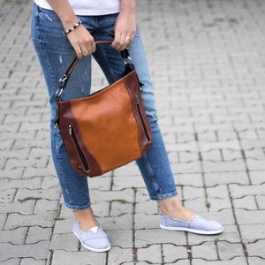 Puede incluir: Una mujer lleva un bolso de cuero marr&oacute;n con detalles negros y una cremallera plateada. El bolso tiene una correa larga y se lleva al hombro. Lleva pantalones vaqueros azules y zapatos de lona grises.