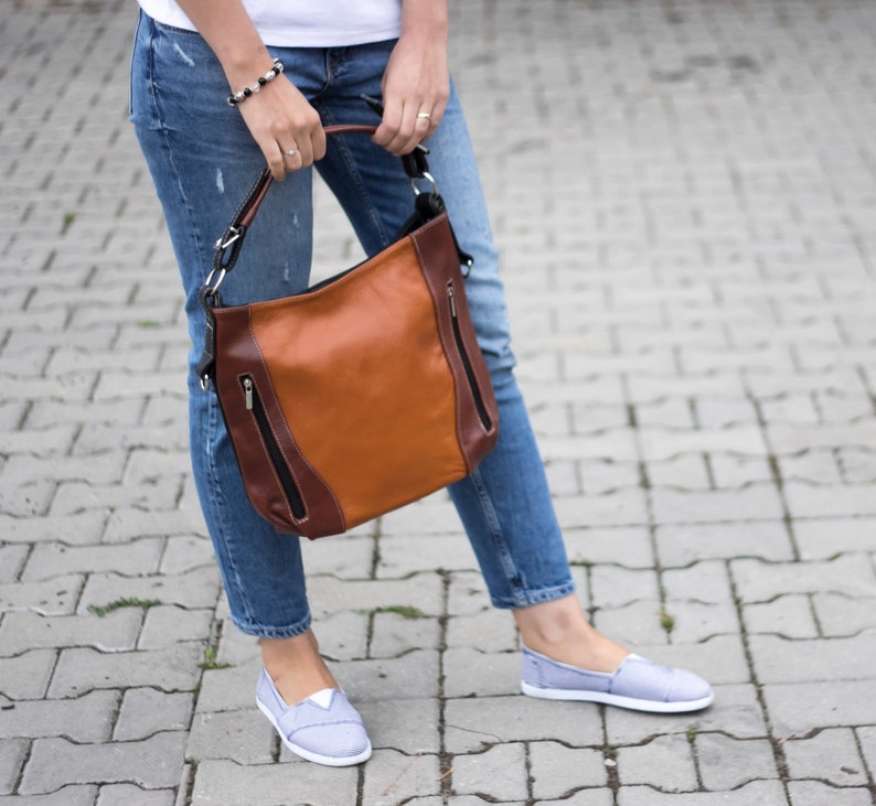 Puede incluir: Una mujer lleva un bolso de cuero marr&oacute;n con detalles negros y una cremallera plateada. El bolso tiene una correa larga y se lleva al hombro. Lleva pantalones vaqueros azules y zapatos de lona grises.
