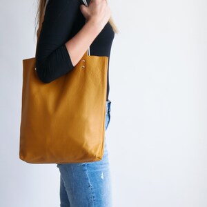 May include: A mustard yellow leather tote bag with white straps. The bag is being carried by a person wearing blue jeans.