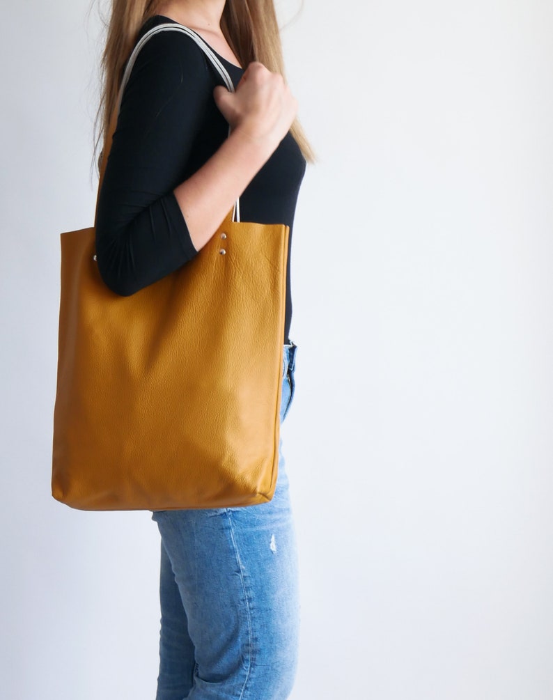 May include: A mustard yellow leather tote bag with white straps. The bag is being carried by a person wearing blue jeans.