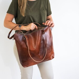 May include: A large, structured brown leather tote bag with a smooth, polished finish. The bag features two thin, light-colored leather shoulder straps, a top zipper closure, and silver-toned metal hardware attachments on the sides.