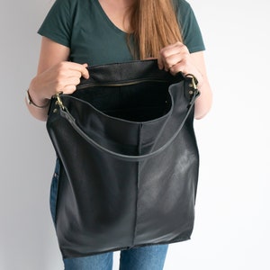 K&ouml;nnte beinhalten: Eine gro&szlig;e schwarze Ledertasche mit Rei&szlig;verschluss und langem Trageriemen. Die Tasche ist offen und das Innere ist sichtbar.