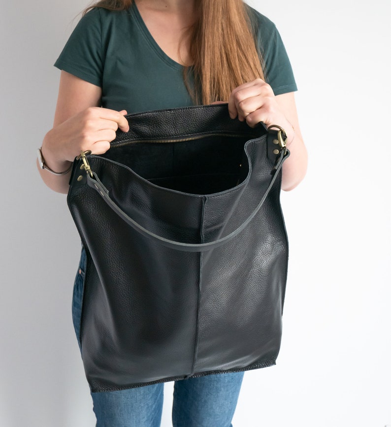 K&ouml;nnte beinhalten: Eine gro&szlig;e schwarze Ledertasche mit Rei&szlig;verschluss und langem Trageriemen. Die Tasche ist offen und das Innere ist sichtbar.