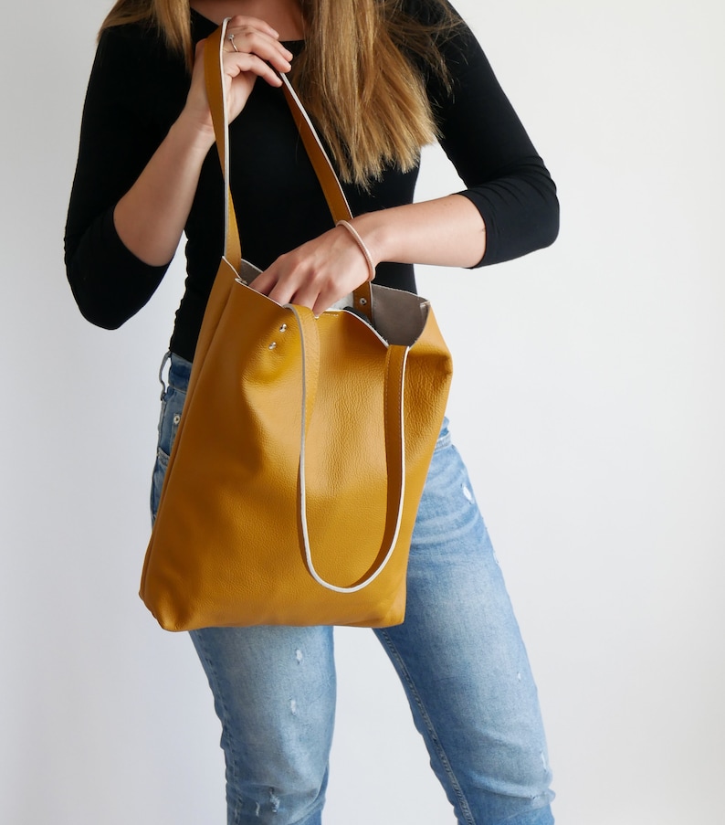 May include: A mustard yellow leather tote bag with white trim and silver hardware. The bag has a wide, flat base and two long straps.