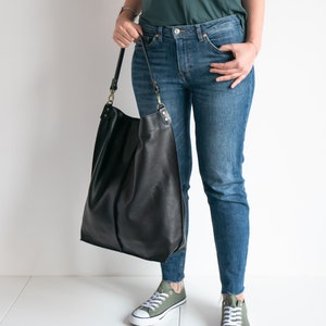 K&ouml;nnte beinhalten: Eine Frau tr&auml;gt eine blaue Jeans und ein dunkelgr&uuml;nes T-Shirt. Sie tr&auml;gt eine gro&szlig;e schwarze Ledertasche mit einem langen Riemen.