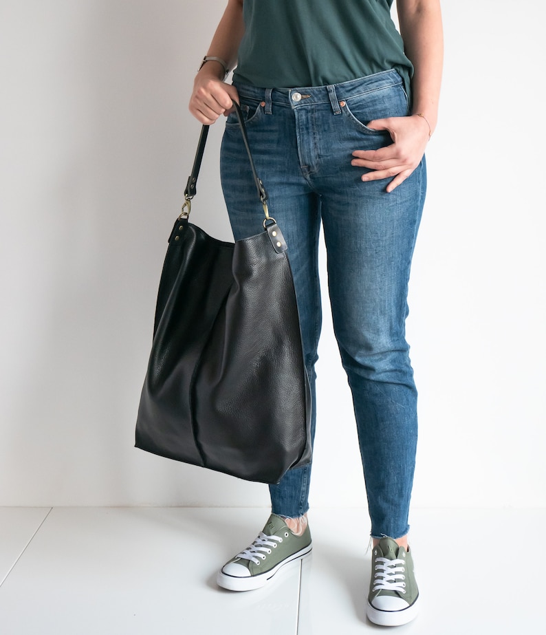 K&ouml;nnte beinhalten: Eine Frau tr&auml;gt eine blaue Jeans und ein dunkelgr&uuml;nes T-Shirt. Sie tr&auml;gt eine gro&szlig;e schwarze Ledertasche mit einem langen Riemen.