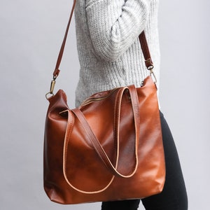 May include: A large, cognac-brown leather tote bag featuring a smooth, polished finish, a top zipper closure, two sturdy shoulder straps, and a detachable, adjustable crossbody strap with antique brass-toned metal hardware clips.
