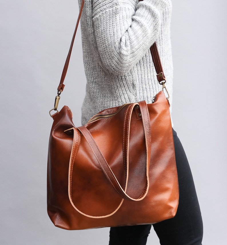May include: A large, cognac-brown leather tote bag featuring a smooth, polished finish, a top zipper closure, two sturdy shoulder straps, and a detachable, adjustable crossbody strap with antique brass-toned metal hardware clips.
