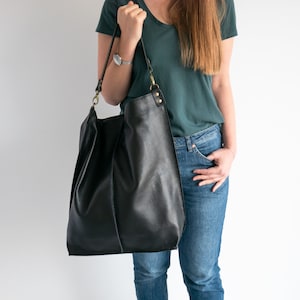 K&ouml;nnte beinhalten: Eine schwarze Ledertasche mit einem langen Trageriemen und goldenen Beschl&auml;gen. Die Tasche wird von einer Person getragen, die ein t&uuml;rkisfarbenes Hemd und eine blaue Jeans tr&auml;gt.