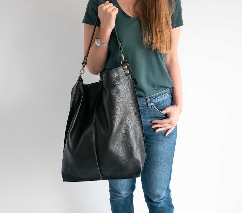 K&ouml;nnte beinhalten: Eine schwarze Ledertasche mit einem langen Trageriemen und goldenen Beschl&auml;gen. Die Tasche wird von einer Person getragen, die ein t&uuml;rkisfarbenes Hemd und eine blaue Jeans tr&auml;gt.