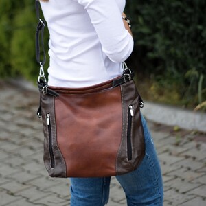 Pode incluir: Uma bolsa de ombro de couro marrom com detalhes em couro preto e z&iacute;peres prateados. A bolsa tem uma al&ccedil;a longa e &eacute; usada no ombro.