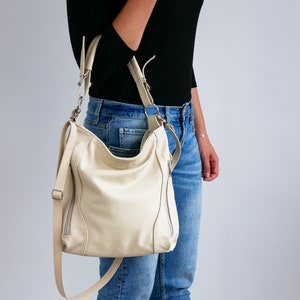 Peut inclure: Un sac &agrave; main en cuir de couleur cr&egrave;me avec une poche zipp&eacute;e de chaque c&ocirc;t&eacute;. Le sac est port&eacute; par une personne v&ecirc;tue d'un jean bleu et d'un chemisier noir.