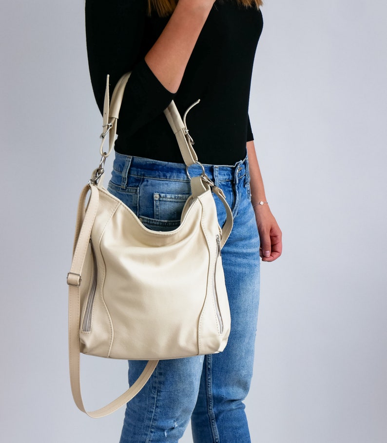 Peut inclure: Un sac &agrave; main en cuir de couleur cr&egrave;me avec une poche zipp&eacute;e de chaque c&ocirc;t&eacute;. Le sac est port&eacute; par une personne v&ecirc;tue d'un jean bleu et d'un chemisier noir.