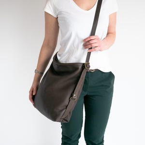 May include: A dark brown leather hobo bag with a long adjustable shoulder strap and antique brass-toned metal hardware. The bag features a slouchy, unstructured silhouette with a wide opening and a single strap attached to the sides with metal rings.