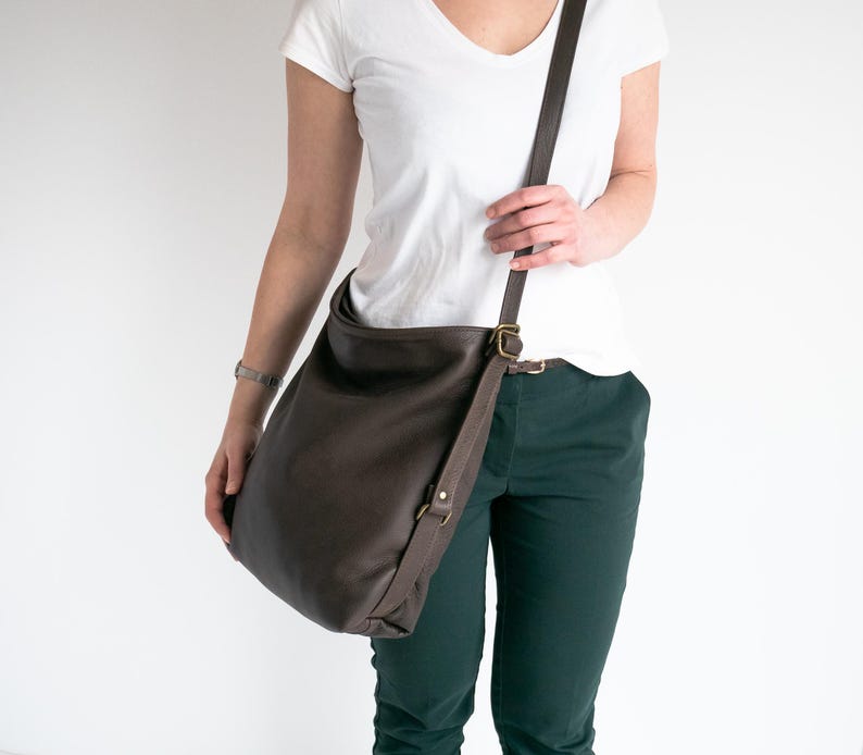 May include: A dark brown leather hobo bag with a long adjustable shoulder strap and antique brass-toned metal hardware. The bag features a slouchy, unstructured silhouette with a wide opening and a single strap attached to the sides with metal rings.