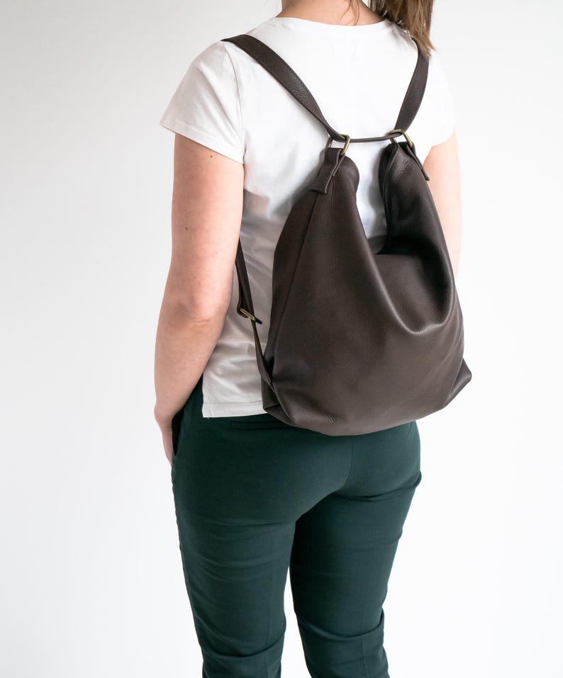 May include: A dark brown leather backpack with a slouchy, unstructured silhouette. The bag features thin shoulder straps attached to metal rings at the top, allowing for a flexible carry. The material has a smooth, matte finish.