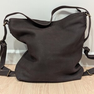 May include: A dark brown leather hobo bag with a pebbled texture, featuring an adjustable shoulder strap and antique brass-toned metal hardware. The bag has a slouchy, unstructured shape with a single main compartment and side strap attachments.