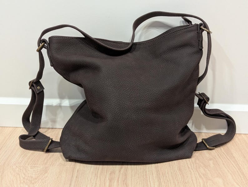May include: A dark brown leather hobo bag with a pebbled texture, featuring an adjustable shoulder strap and antique brass-toned metal hardware. The bag has a slouchy, unstructured shape with a single main compartment and side strap attachments.