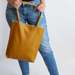 May include: A mustard yellow leather tote bag with white trim and silver hardware. The bag is being held by a person wearing blue jeans.