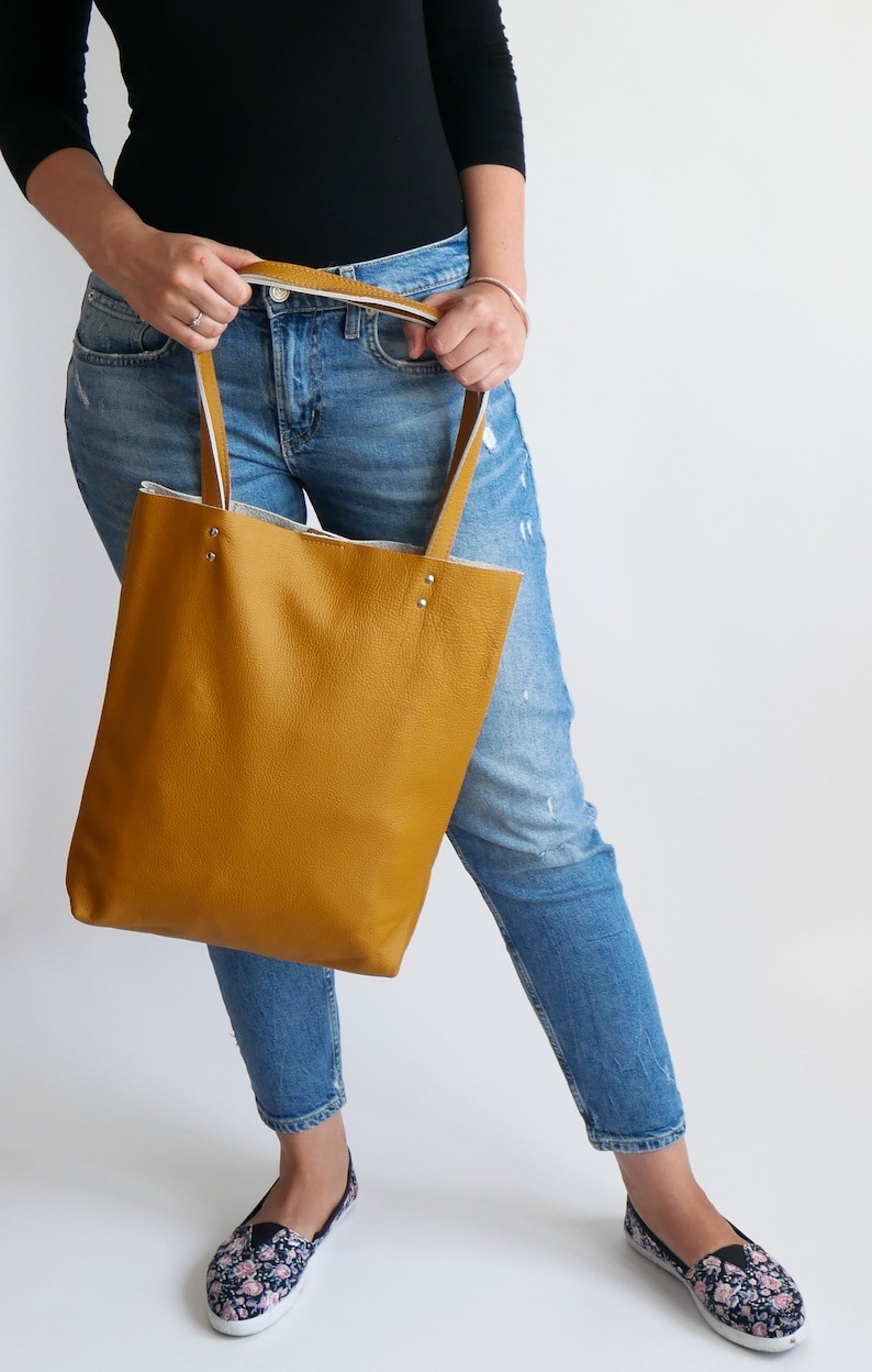 May include: A mustard yellow leather tote bag with white trim and silver hardware. The bag is being held by a person wearing blue jeans.