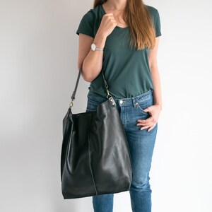 K&ouml;nnte beinhalten: Eine Frau tr&auml;gt ein t&uuml;rkisfarbenes T-Shirt und eine blaue Jeans. Sie tr&auml;gt eine gro&szlig;e schwarze Ledertasche mit einem braunen Riemen.