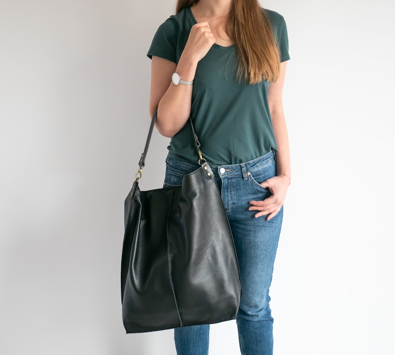 K&ouml;nnte beinhalten: Eine Frau tr&auml;gt ein t&uuml;rkisfarbenes T-Shirt und eine blaue Jeans. Sie tr&auml;gt eine gro&szlig;e schwarze Ledertasche mit einem braunen Riemen.