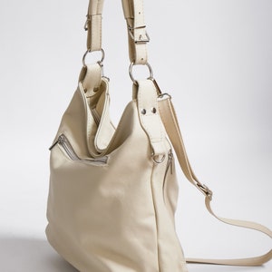 Peut inclure: Un sac &agrave; main en cuir de couleur cr&egrave;me avec une longue bandouli&egrave;re pouvant &ecirc;tre port&eacute; comme un sac &agrave; dos ou un sac &agrave; bandouli&egrave;re. Le sac a plusieurs poches et une fermeture &agrave; glissi&egrave;re.