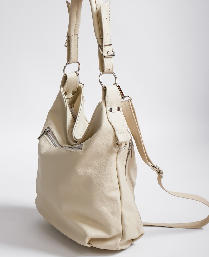 Peut inclure: Un sac &agrave; main en cuir de couleur cr&egrave;me avec une longue bandouli&egrave;re pouvant &ecirc;tre port&eacute; comme un sac &agrave; dos ou un sac &agrave; bandouli&egrave;re. Le sac a plusieurs poches et une fermeture &agrave; glissi&egrave;re.