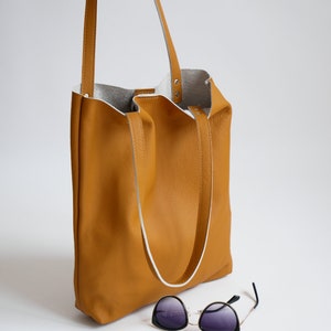 May include: A mustard yellow leather tote bag with white leather straps and a white leather lining. The bag is sitting on a white surface with a pair of black sunglasses in front of it.
