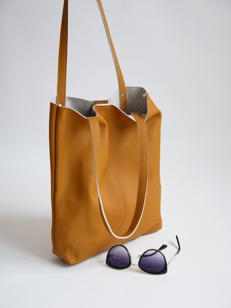 May include: A mustard yellow leather tote bag with white leather straps and a white leather lining. The bag is sitting on a white surface with a pair of black sunglasses in front of it.