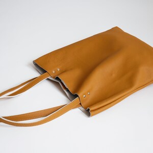 May include: A brown leather tote bag with white leather straps. The bag is lying on a white surface.