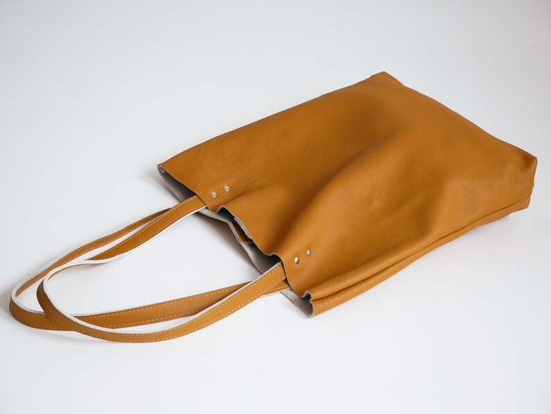 May include: A brown leather tote bag with white leather straps. The bag is lying on a white surface.