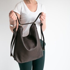 May include: A dark brown leather backpack with a slouchy, unstructured silhouette. The bag features thin adjustable shoulder straps attached with antique brass-toned metal hardware, including rectangular rings and sliders for length adjustment.