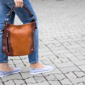 Puede incluir: Un bolso de cuero marr&oacute;n con ribetes marrones oscuros y herrajes plateados. El bolso tiene dos compartimentos con cremallera y un asa superior.