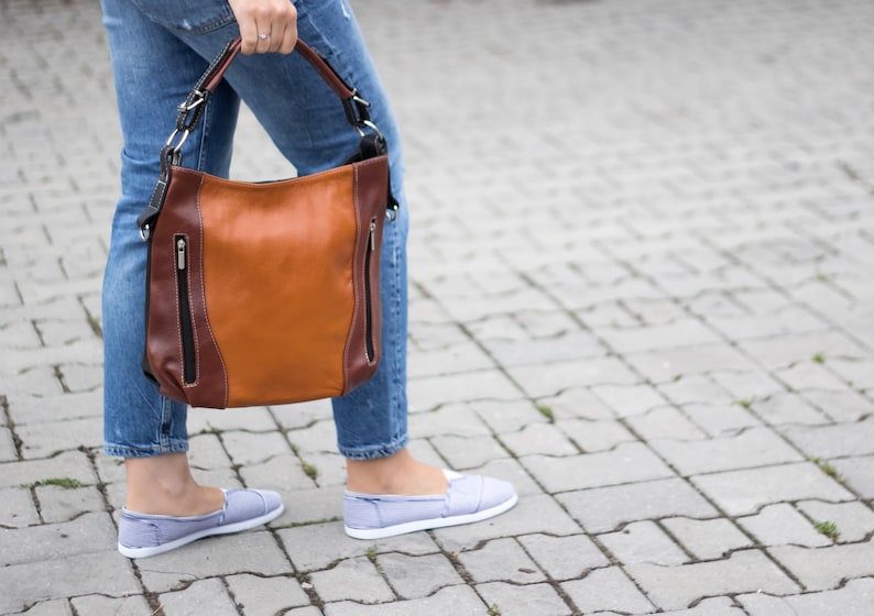 Puede incluir: Un bolso de cuero marr&oacute;n con ribetes marrones oscuros y herrajes plateados. El bolso tiene dos compartimentos con cremallera y un asa superior.