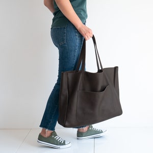 May include: A large, dark brown leather tote bag with a front pocket and long handles. The bag is held by a person wearing blue jeans and green sneakers. The bag appears to be empty.