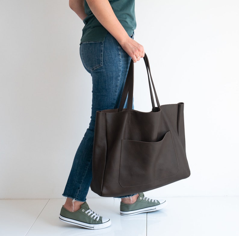 May include: A large, dark brown leather tote bag with a front pocket and long handles. The bag is held by a person wearing blue jeans and green sneakers. The bag appears to be empty.