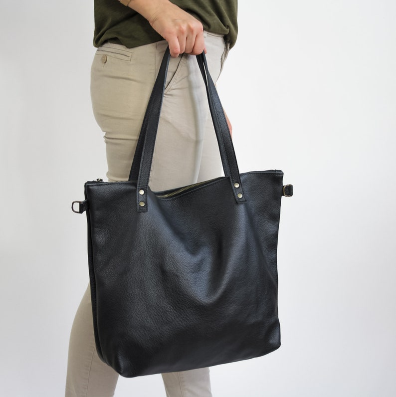 May include: A black tote bag with a textured surface, featuring two long shoulder straps attached with metallic rivets, a top zipper closure, and side hardware loops for additional attachments on a plain background.