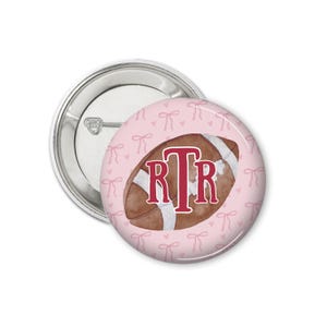 May include: A pink button with a white and brown football with the monogram "RTR" in red. The background is a pink pattern of small hearts and bows.