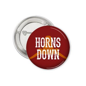 May include: A red button with the text "HORNS DOWN" in white lettering. The text is in a distressed font. A gold silhouette of a pair of horns is behind the text.