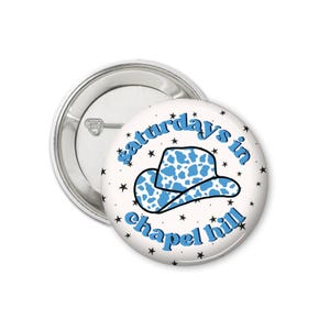 Peut inclure: Un badge blanc avec un chapeau de cowboy bleu et le texte "saturdays in chapel hill" en bleu. Le chapeau a un motif de vache. De petites &eacute;toiles noires entourent le design.
